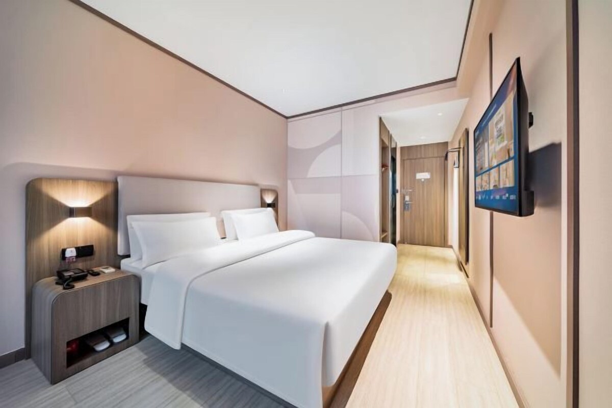 A modern hotel room with a large bed, bedside tables, a TV, and a hallway leading to another room.