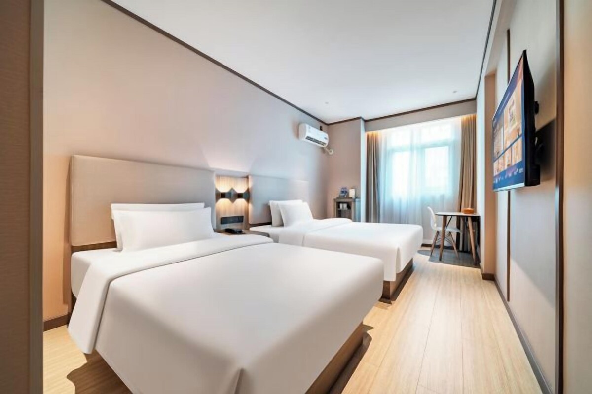 A hotel room with two beds, a TV, a desk, and a window with curtains.