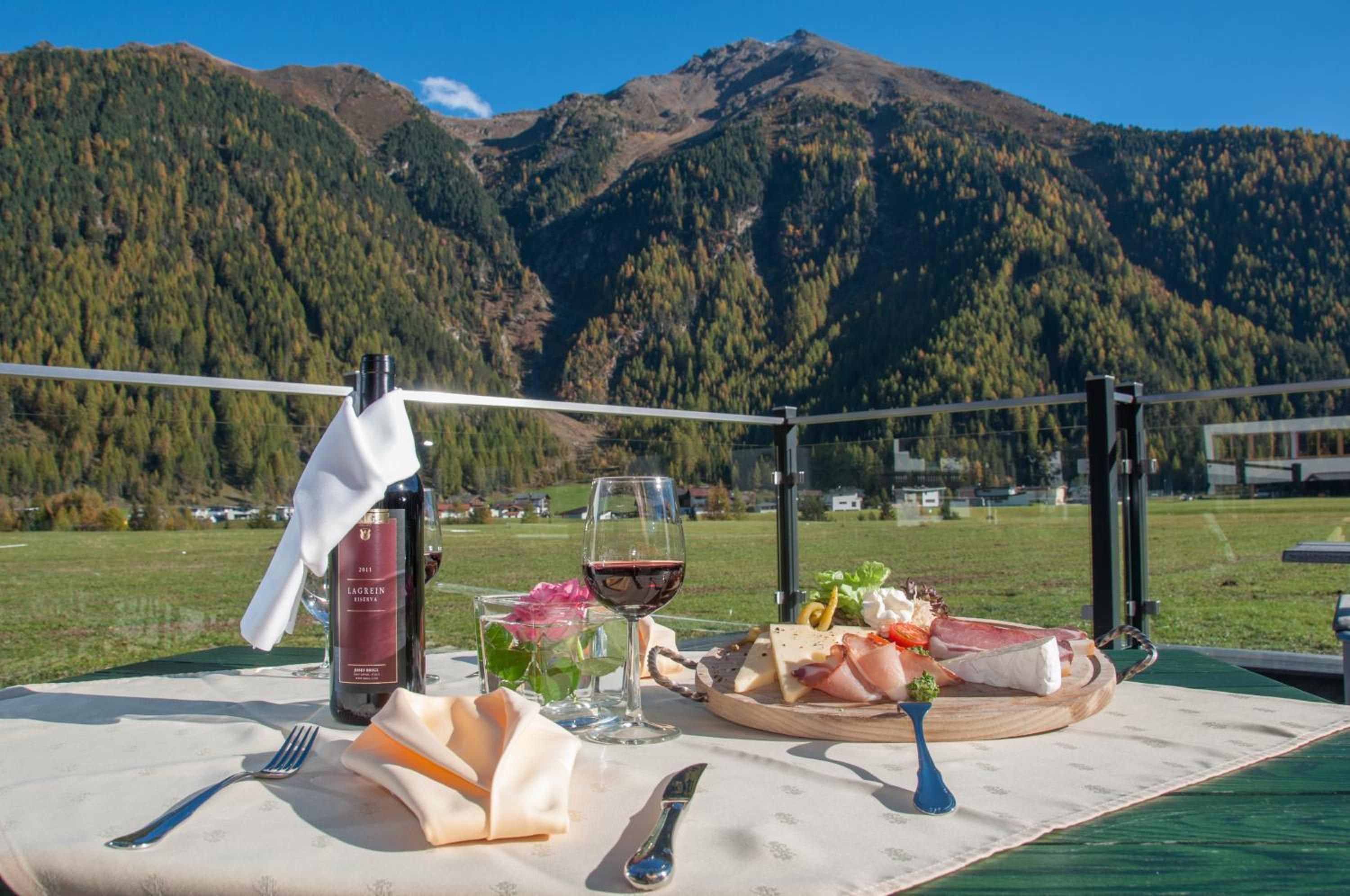 A dining table set outdoors with a bottle, glasses, and a plate of food against a mountain backdrop.