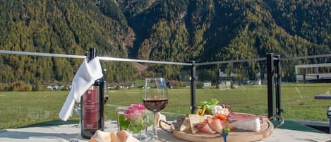 A dining table set outdoors with a bottle, glasses, and a plate of food against a mountain backdrop.