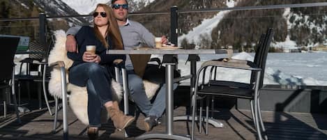 A couple seated at an outdoor table with a mountain view, surrounded by chairs and a wooden deck.