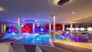 A modern indoor swimming pool area with multiple lanes, a large water slide, and vibrant lighting. - Hotel Seehof (Koessen)