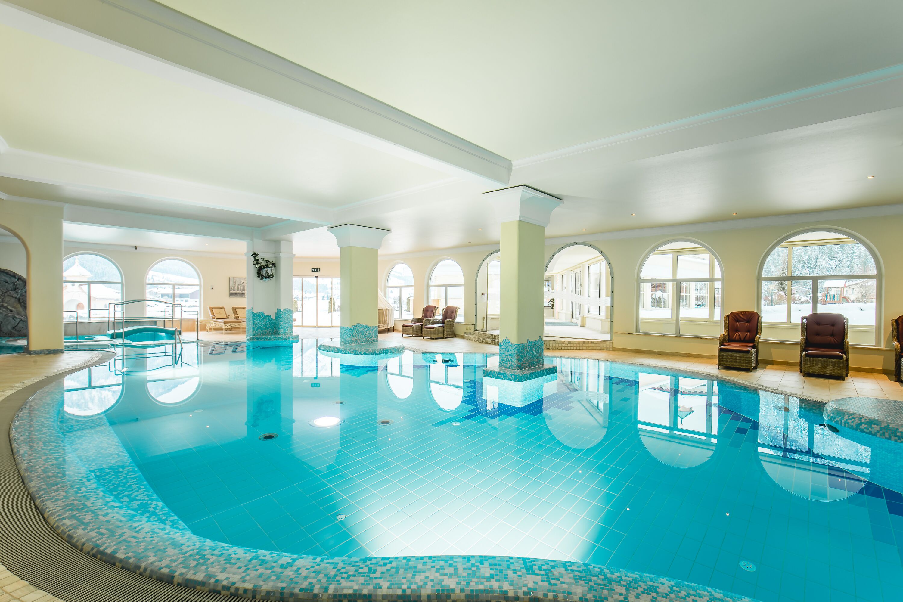 An indoor swimming pool with clear blue water, surrounded by large arched windows and furnished with lounge chairs.
