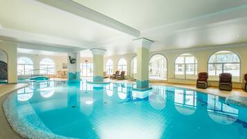 An indoor swimming pool with clear blue water, surrounded by large arched windows and furnished with lounge chairs.