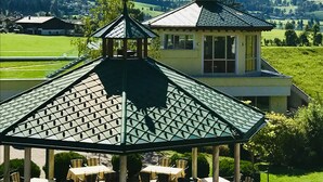 A gazebo with a green roof and a small tower, surrounded by outdoor tables and chairs, set against a backdrop of mountains and a clear sky. - Hotel Seehof (Koessen)