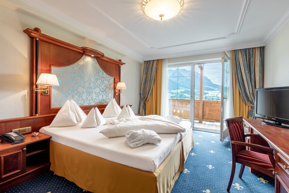 Habitación de hotel con una cama grande, un escritorio, una silla, un televisor y un balcón con vista a las montañas.