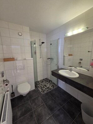Un baño moderno con ducha envuelta en vidrio, inodoro blanco y lavamanos con encimera oscura.