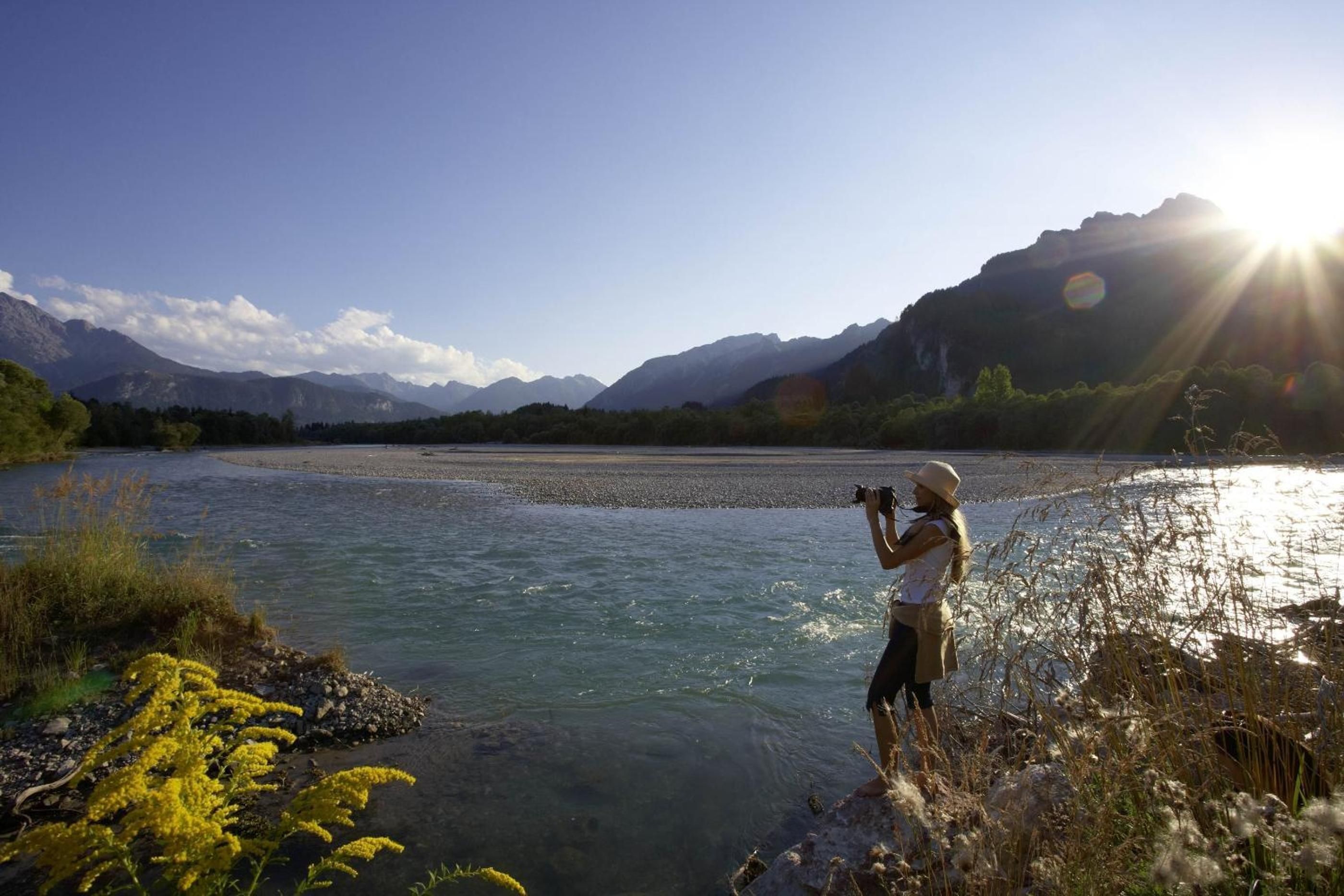 A person taking a photograph of a river with mountains in the background.