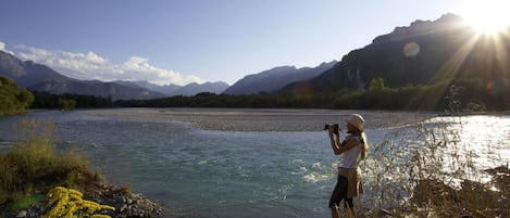 A person taking a photograph of a river with mountains in the background.