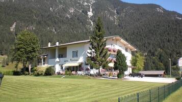 Un hotel con balcón, césped y montañas al fondo.