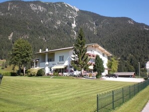 Un hotel con balcón, césped y montañas al fondo.