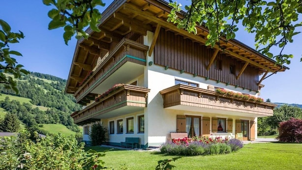 A two-story chalet with wooden balconies, a covered porch, and a lush garden. - Pension Sonnblick (WESTENDORF)
