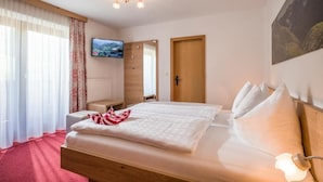 A hotel room with a bed, a TV mounted on the wall, a wooden door, and a window with curtains. - Pension Sonnblick (WESTENDORF)