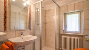 A bathroom with a glass-enclosed shower, a sink, a toilet, and a window with blinds. - Pension Sonnblick (WESTENDORF)