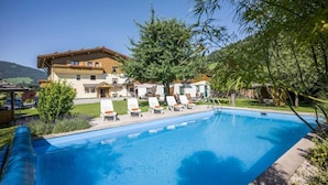 A swimming pool with lounge chairs, a building with a sloped roof, and a tree in the foreground. - Pension Sonnblick (WESTENDORF)