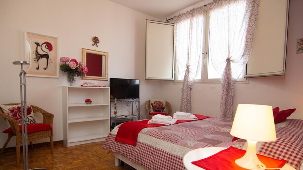 A bedroom with a bed, a chair, a TV, a shelf with a vase, and a window with curtains.