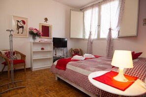 A bedroom with a bed, a chair, a TV, a shelf with a vase, and a window with curtains.