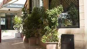 Um edifício com a placa da BancaMarch, plantas em vasos e uma grande janela refletindo o ambiente.