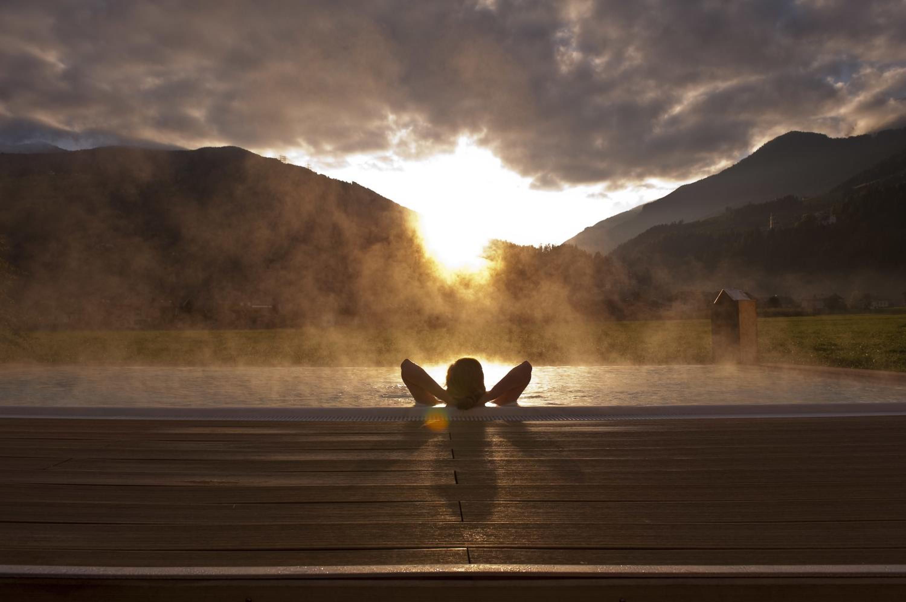 A wooden deck overlooking a hot spring with steam rising, surrounded by mountains and a cloudy sky.