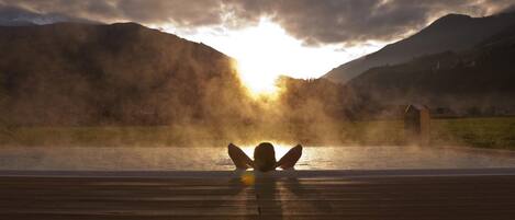 A wooden deck overlooking a hot spring with steam rising, surrounded by mountains and a cloudy sky.