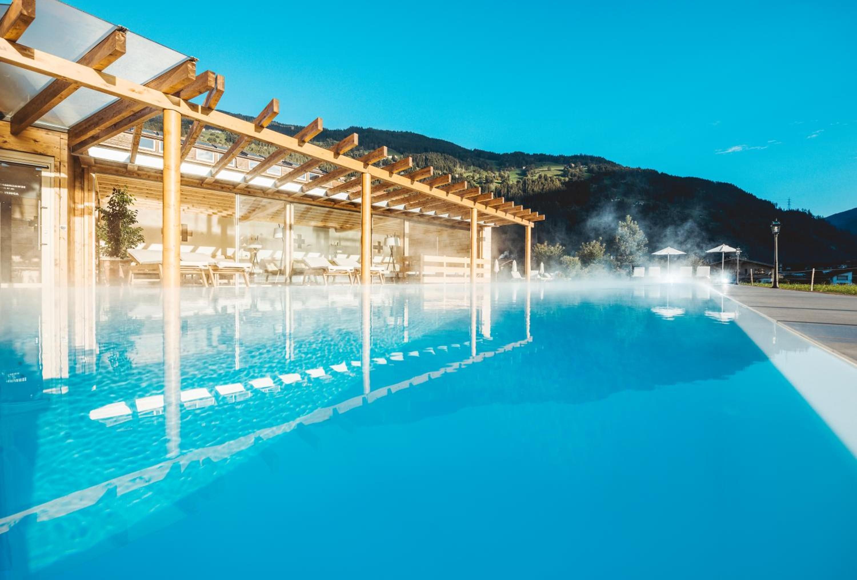 A clear blue pool with wooden pergolas and lounge chairs, surrounded by a mountainous landscape.