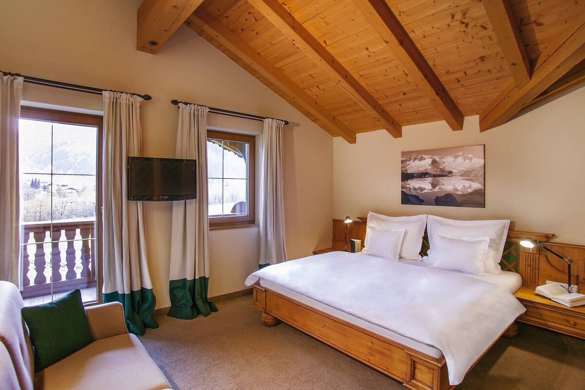 A bedroom with a wooden ceiling, a large bed, a TV mounted on the wall, and a balcony view.