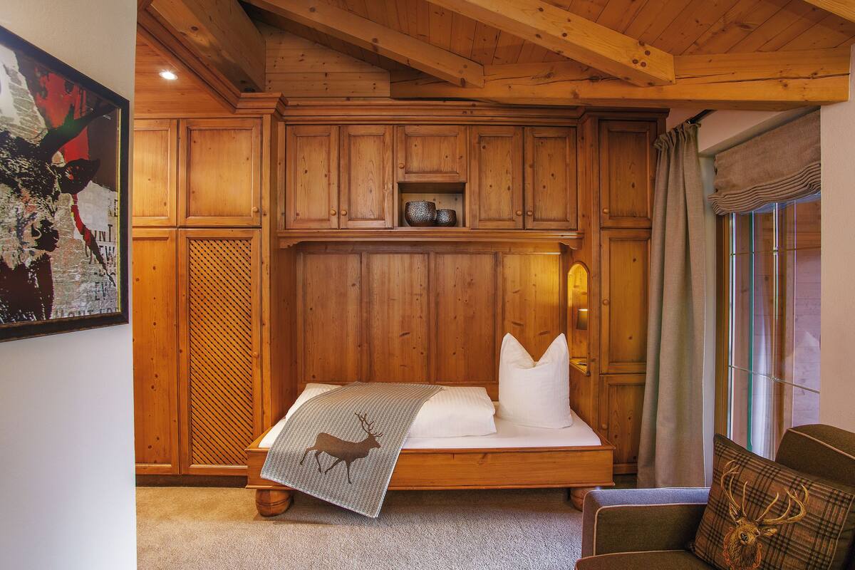 A wooden cabin interior with a bed, a chair, and a window with curtains.