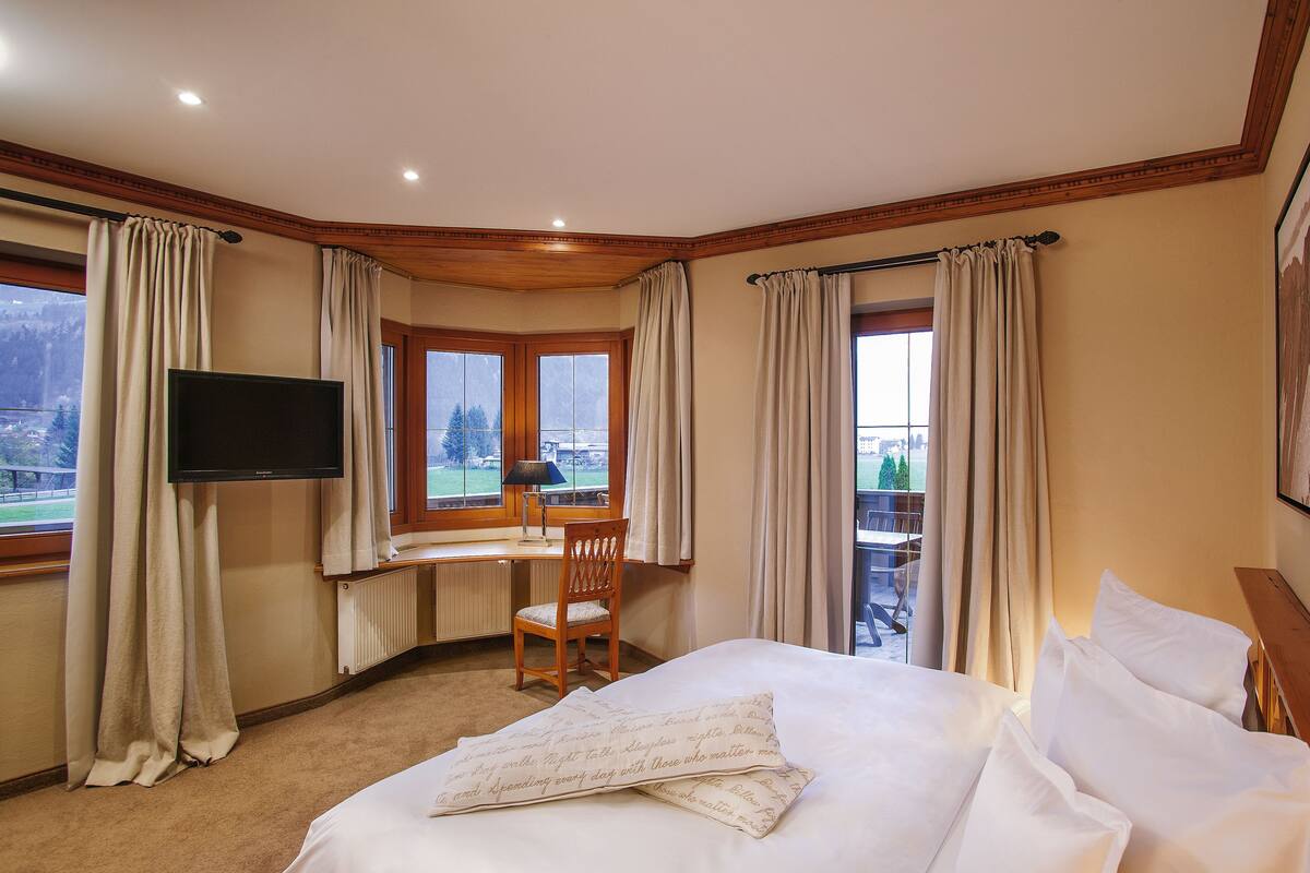 A hotel room with a bed, a desk, a chair, a TV, and large windows with curtains.