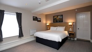 Een slaapkamer met een groot bed, een nachtkastje, een hoofdbord met verlichting, een raam met gordijnen en een deur.