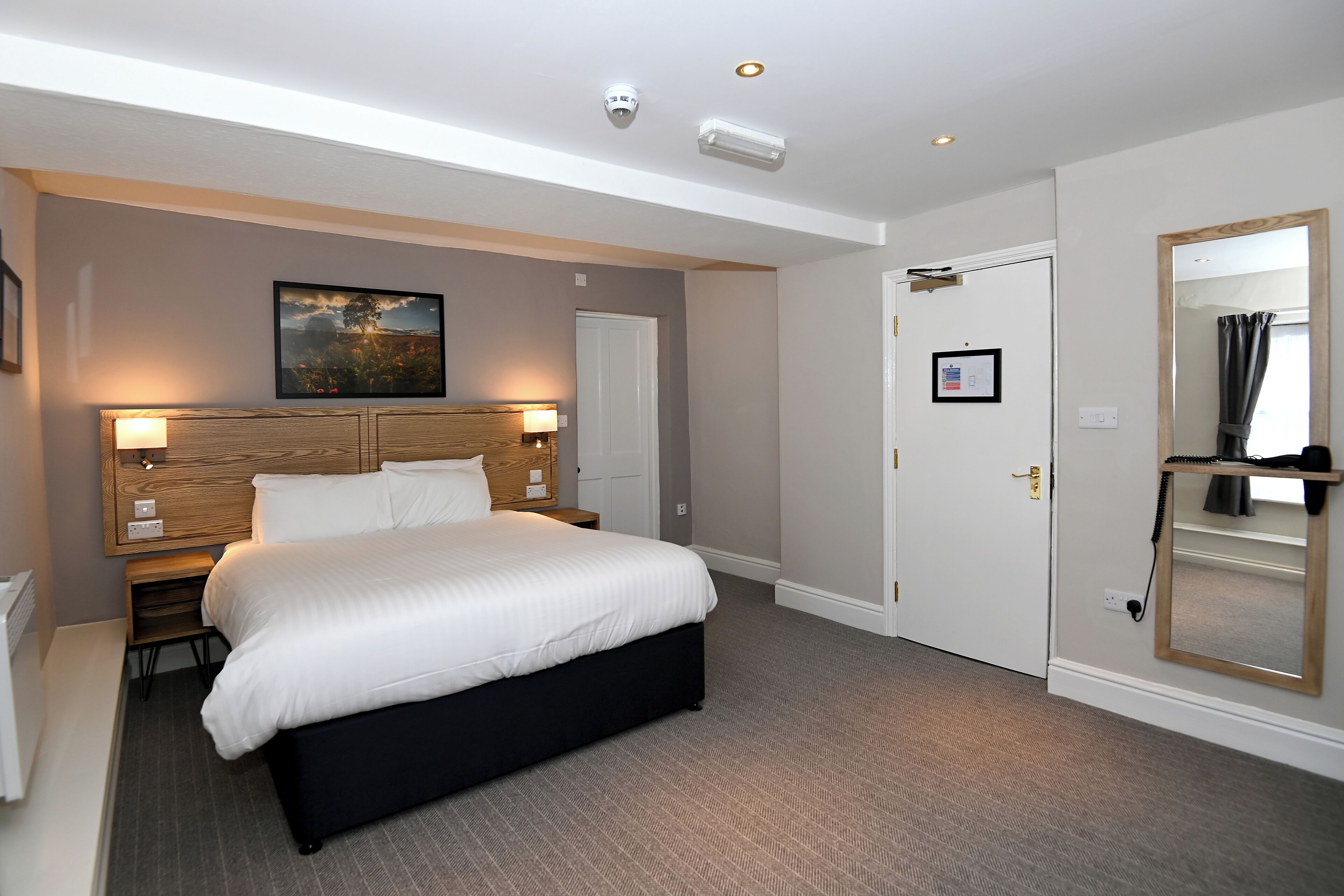 Habitación de hotel con una cama grande, cabecera de madera, mesita de noche, lámpara de pared, cuadro enmarco, puerta con espejo y suelo alfombrado.
