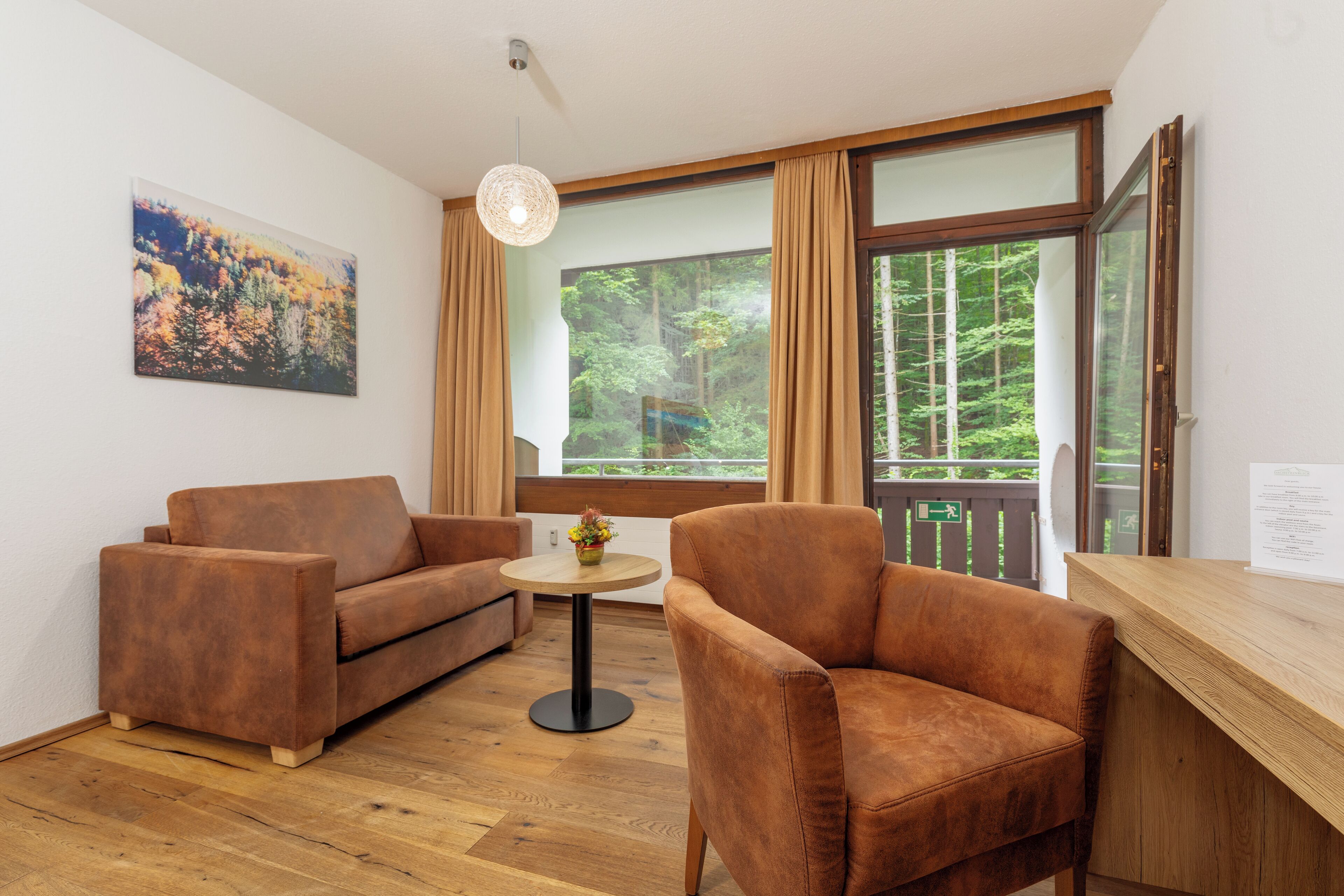 Una habitación con piso de madera, dos sofás marrones, una mesa redonda con un florero y vista a un bosque a través de amplios ventanales.