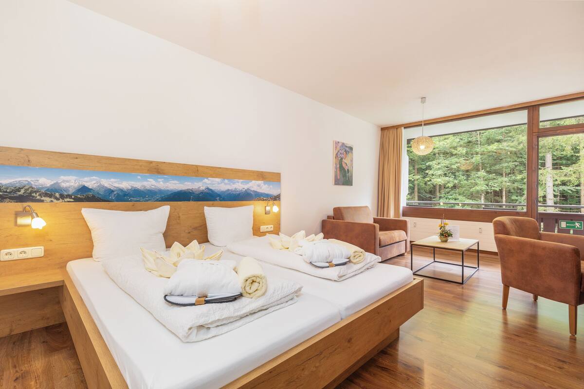 A modern hotel room with a large bed, wooden headboard, and a painting of mountains. There is a seating area with a table and chairs, and a large window with a view of trees.