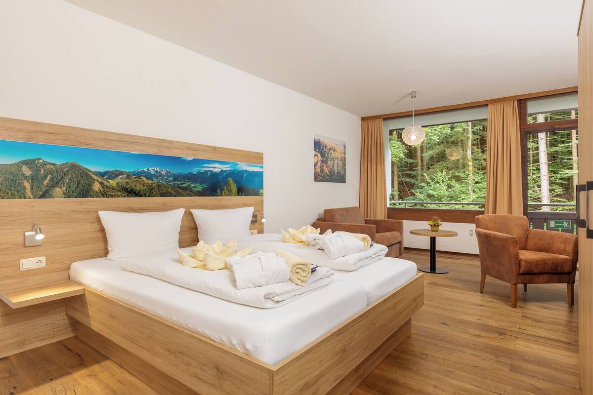 A modern hotel room with a large bed, wooden headboard, and a painting of a mountain landscape. There is a small table with a plant and a chair in the corner.