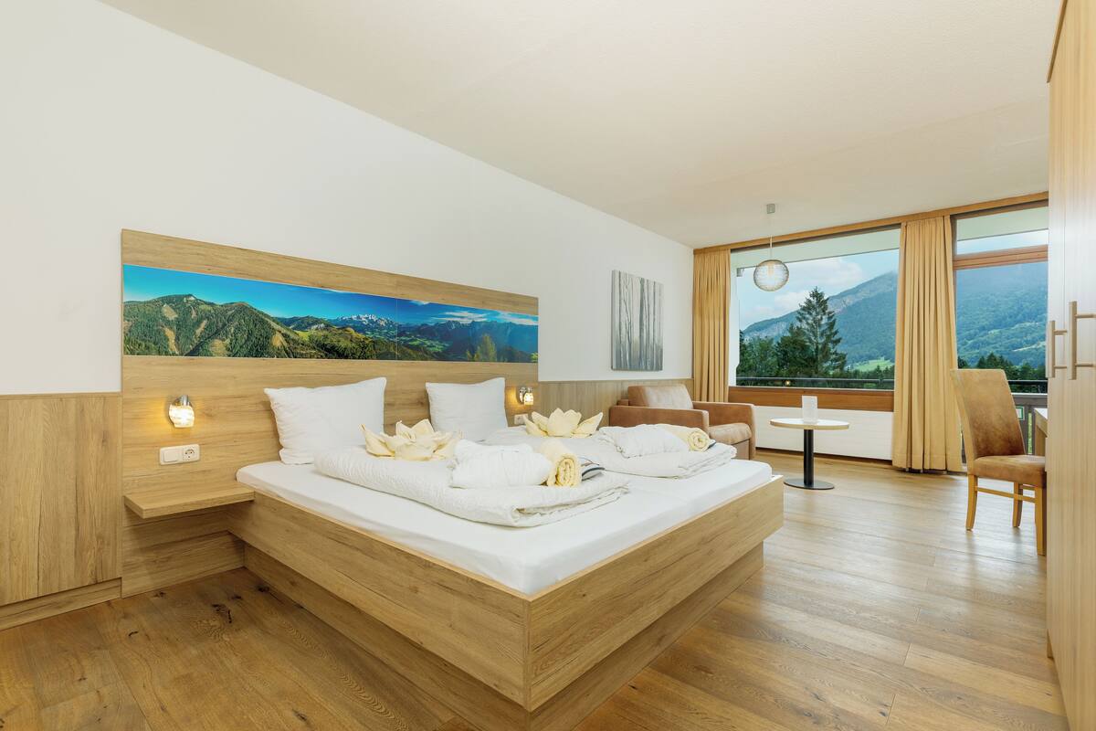 A modern hotel room with a wooden bed, a painting of a mountain landscape, a small table, and a chair.