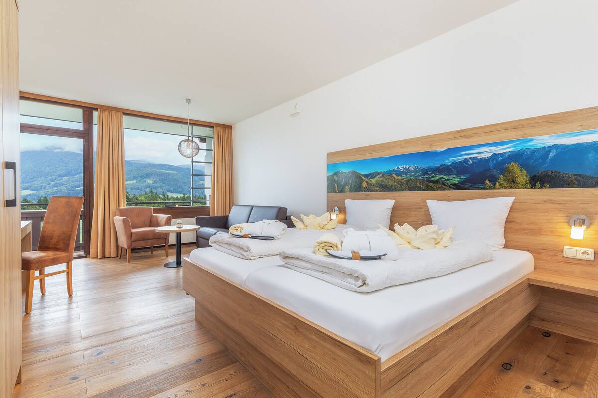 A modern hotel room with a large bed, a desk, a chair, and a view of mountains through large windows.