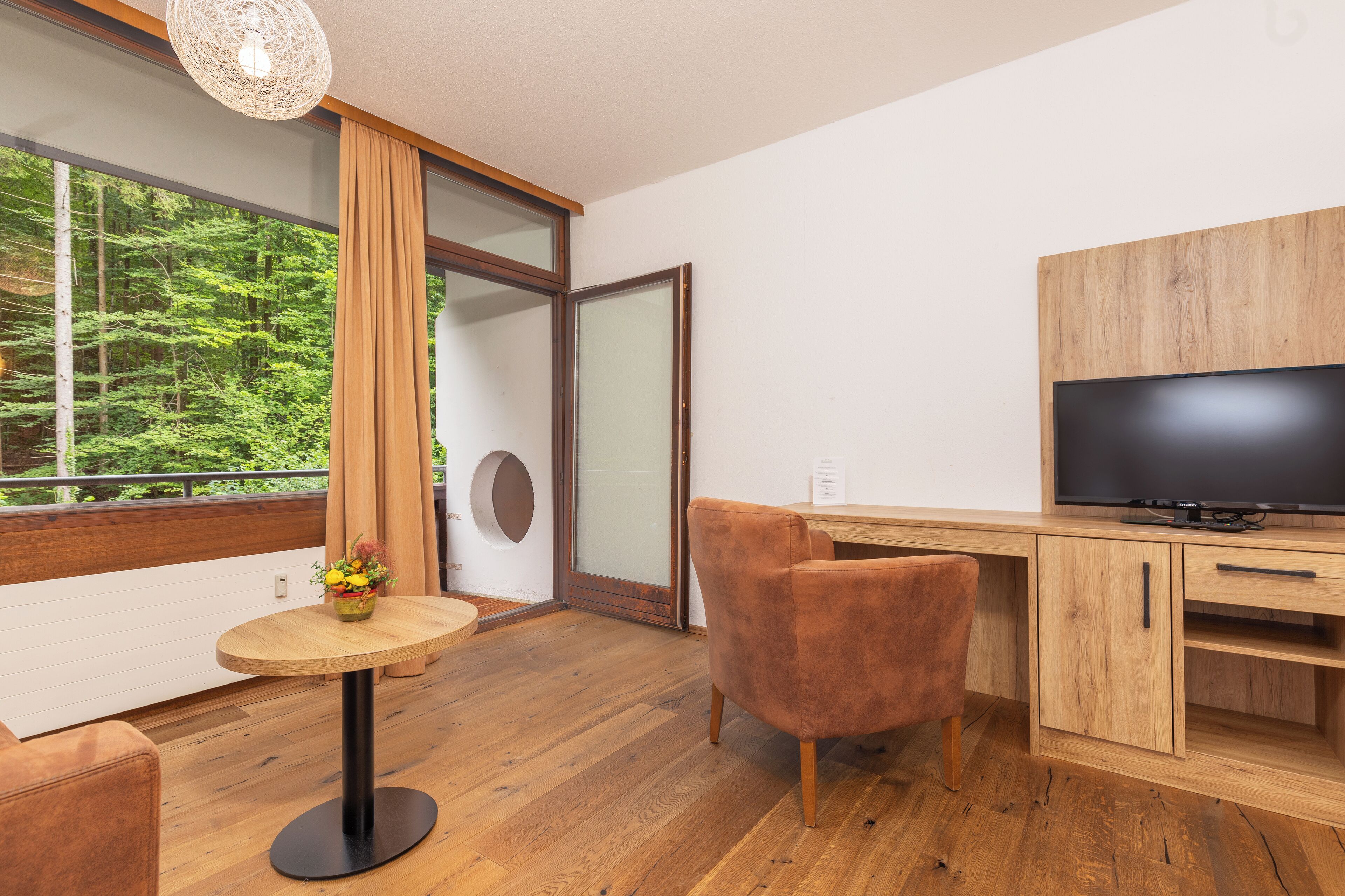A modern room with a wooden floor, a round table with a vase of flowers, a brown chair, a TV on a wooden cabinet, and a large window with a view of trees.