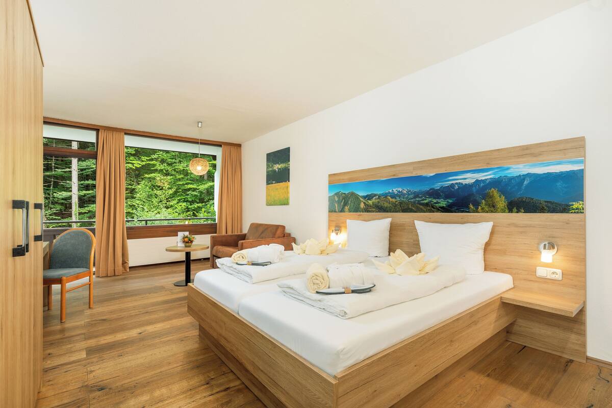 A modern hotel room with a large bed, a wooden headboard, a view of greenery through the window, and a painting of mountains on the wall.