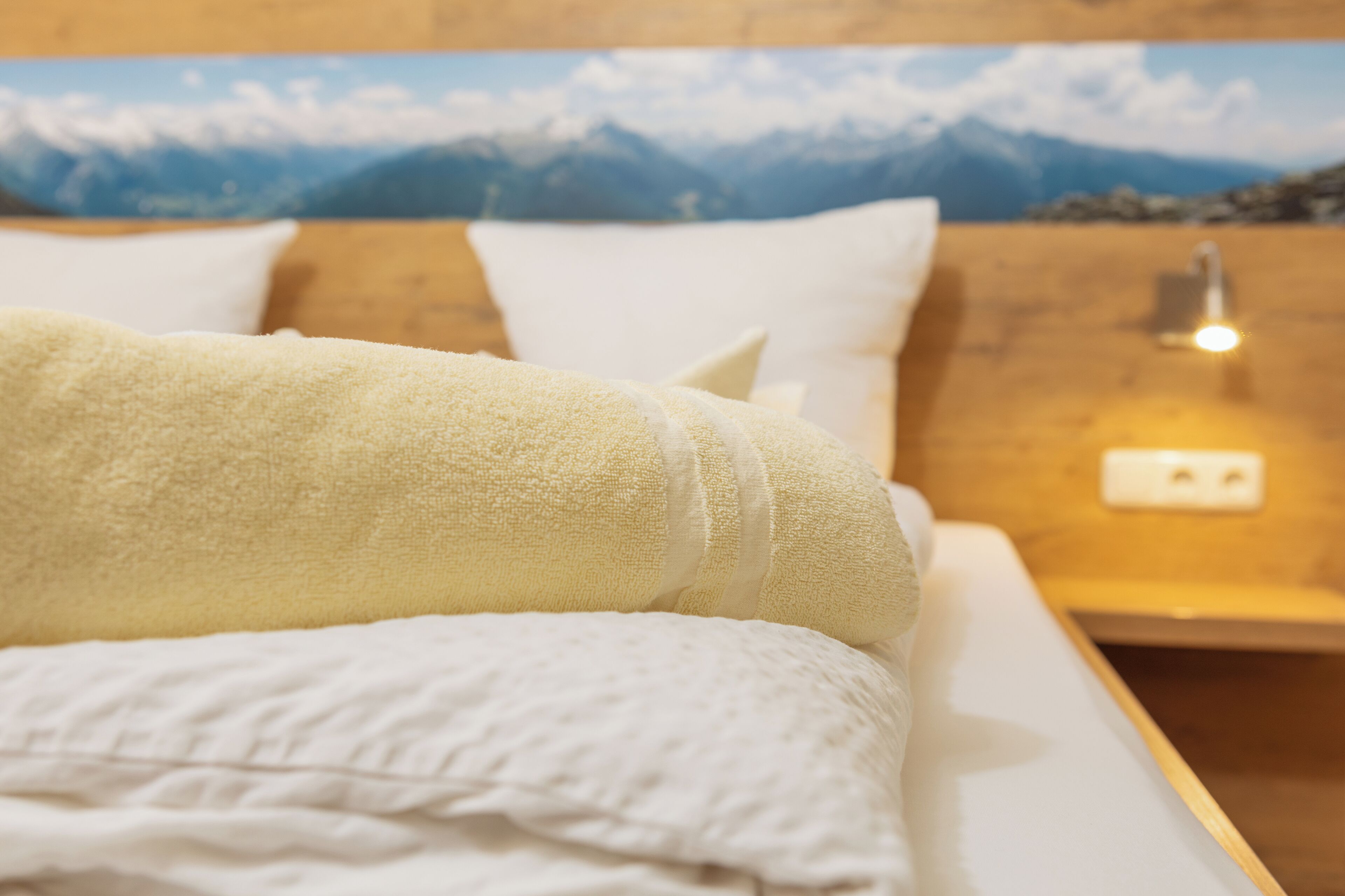 Una cama con ropa blanca y una toalla amarilla, cabecera de madera y una lámpara fijada a la pared.