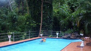 Piscina cercada por vegetação exuberante, com deck de azulejos e grade de metal.