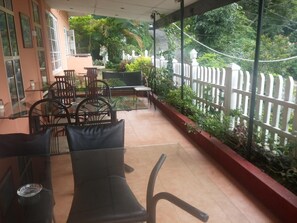A covered outdoor seating area with tables and chairs, surrounded by a white fence and lush greenery.