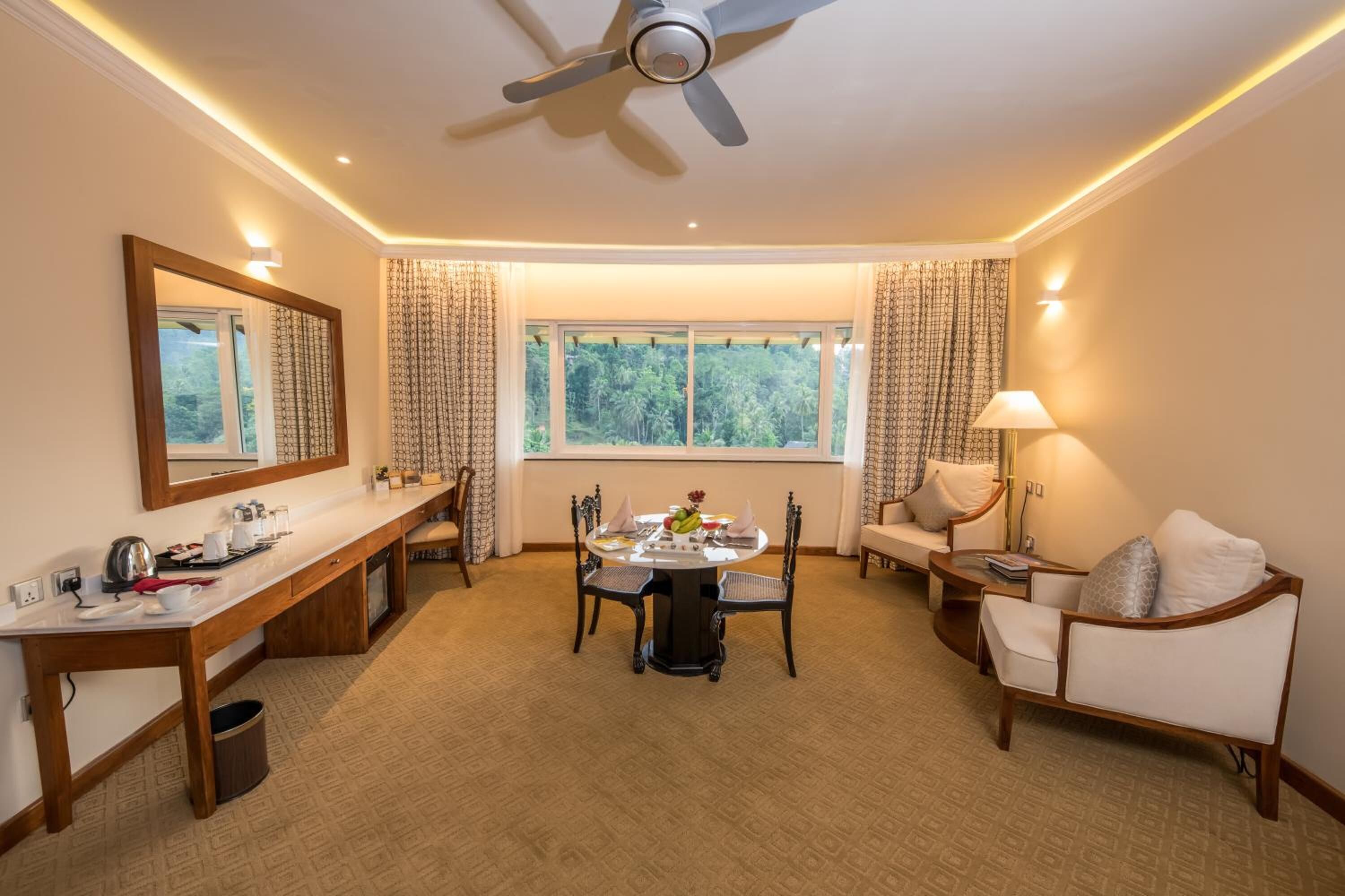 Habitación de hotel con una mesa de comedor, sillas, un sofá, un televisor, un ventilador de techo y una ventana con cortinas.