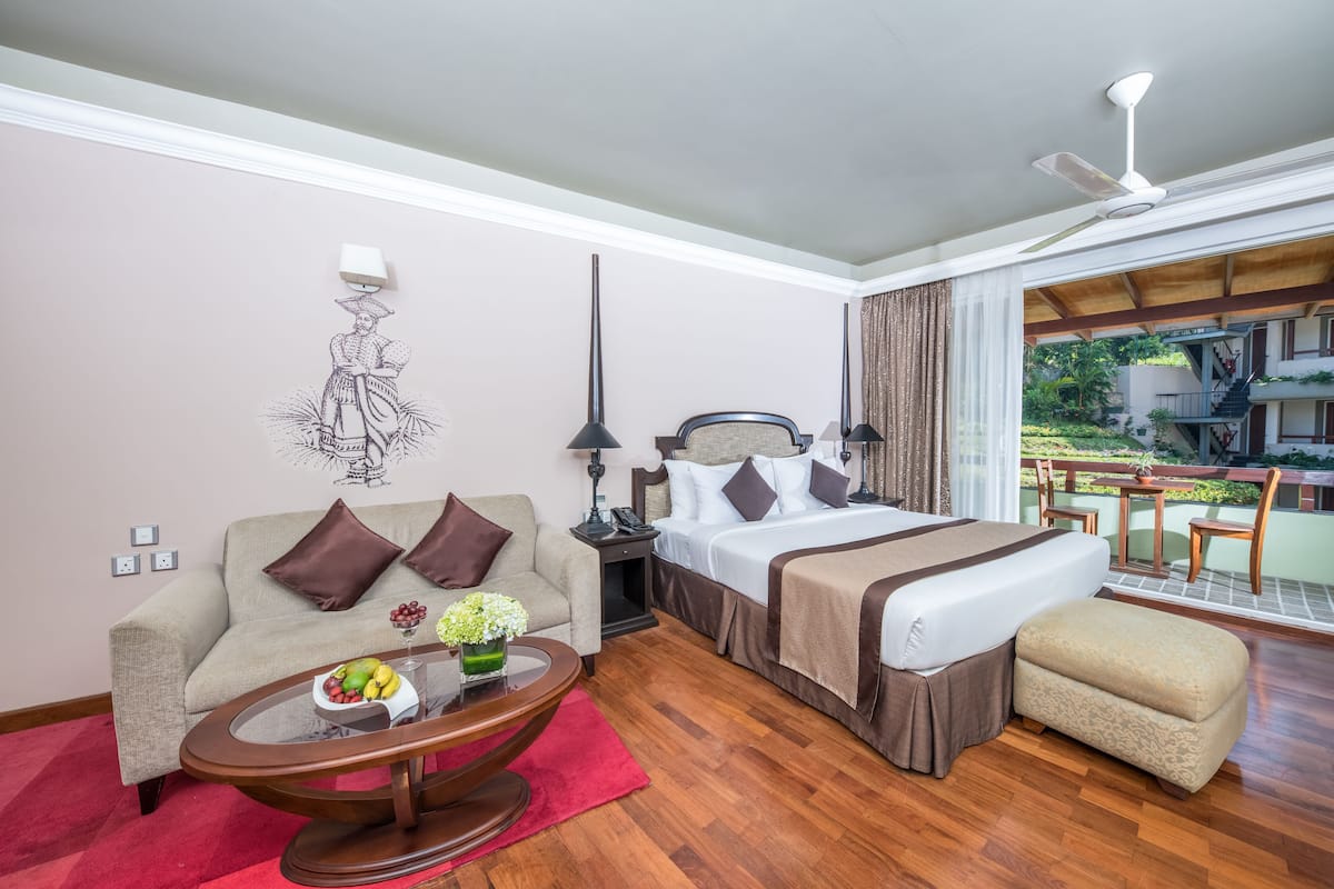 Habitación de hotel con cama, sofá, mesa de centro con fruta, lámpara y vistas al exterior.