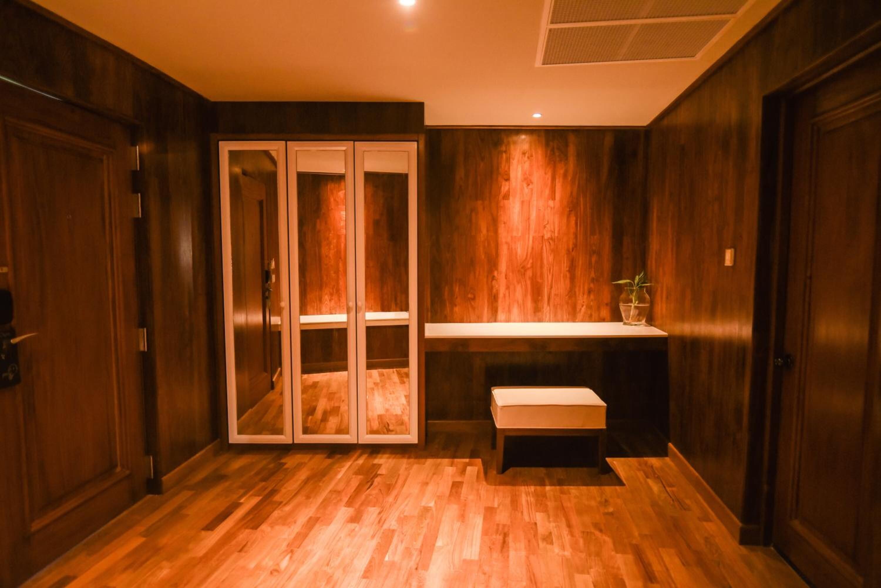 A room with wooden flooring, a bench, a shelf with a plant, and a mirrored cabinet.