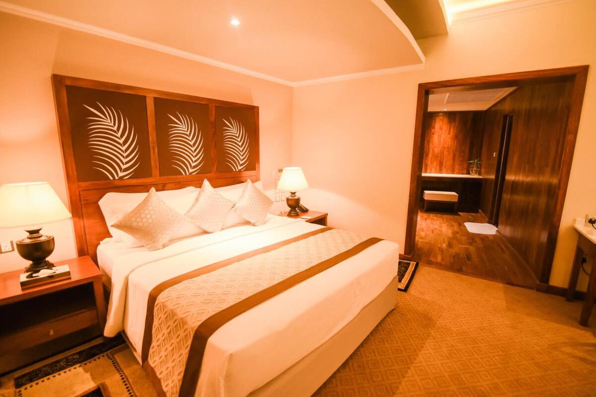 Habitación de hotel con una cama grande, dos mesitas de noche con lámparas, cabecero de madera con diseños de hojas y un baño visible a través de una puerta abierta.