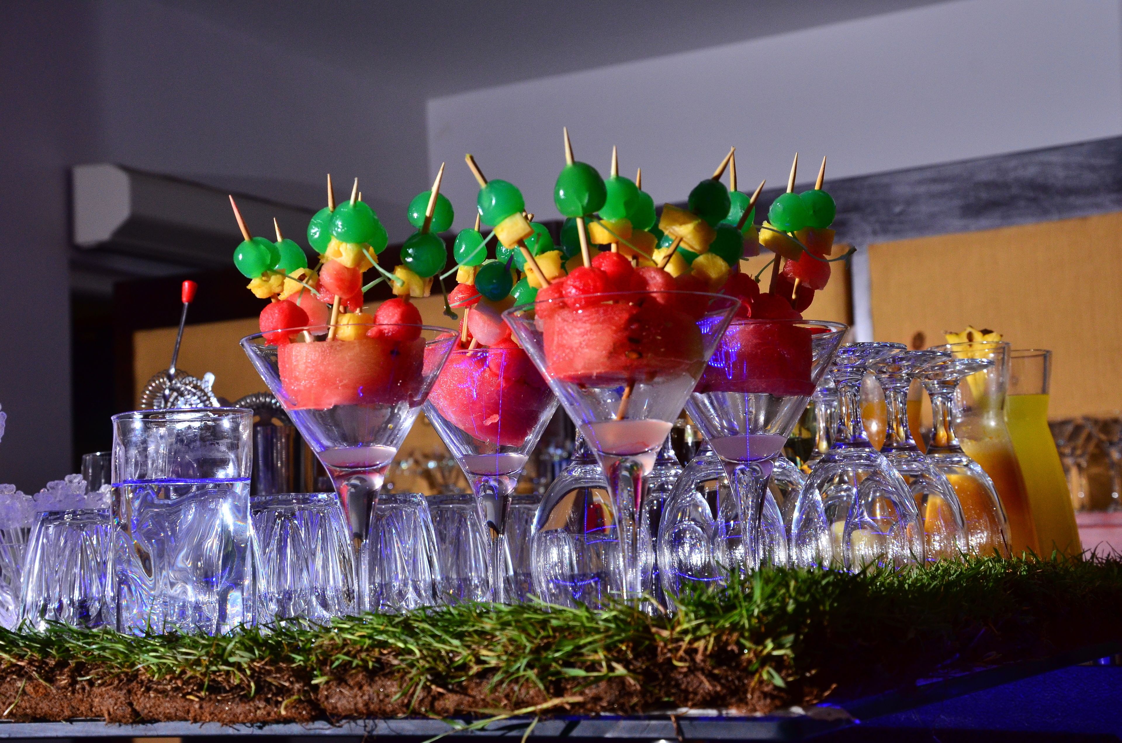 A table with martini glasses filled with watermelon and fruit skewers, surrounded by ice cubes and glasses.