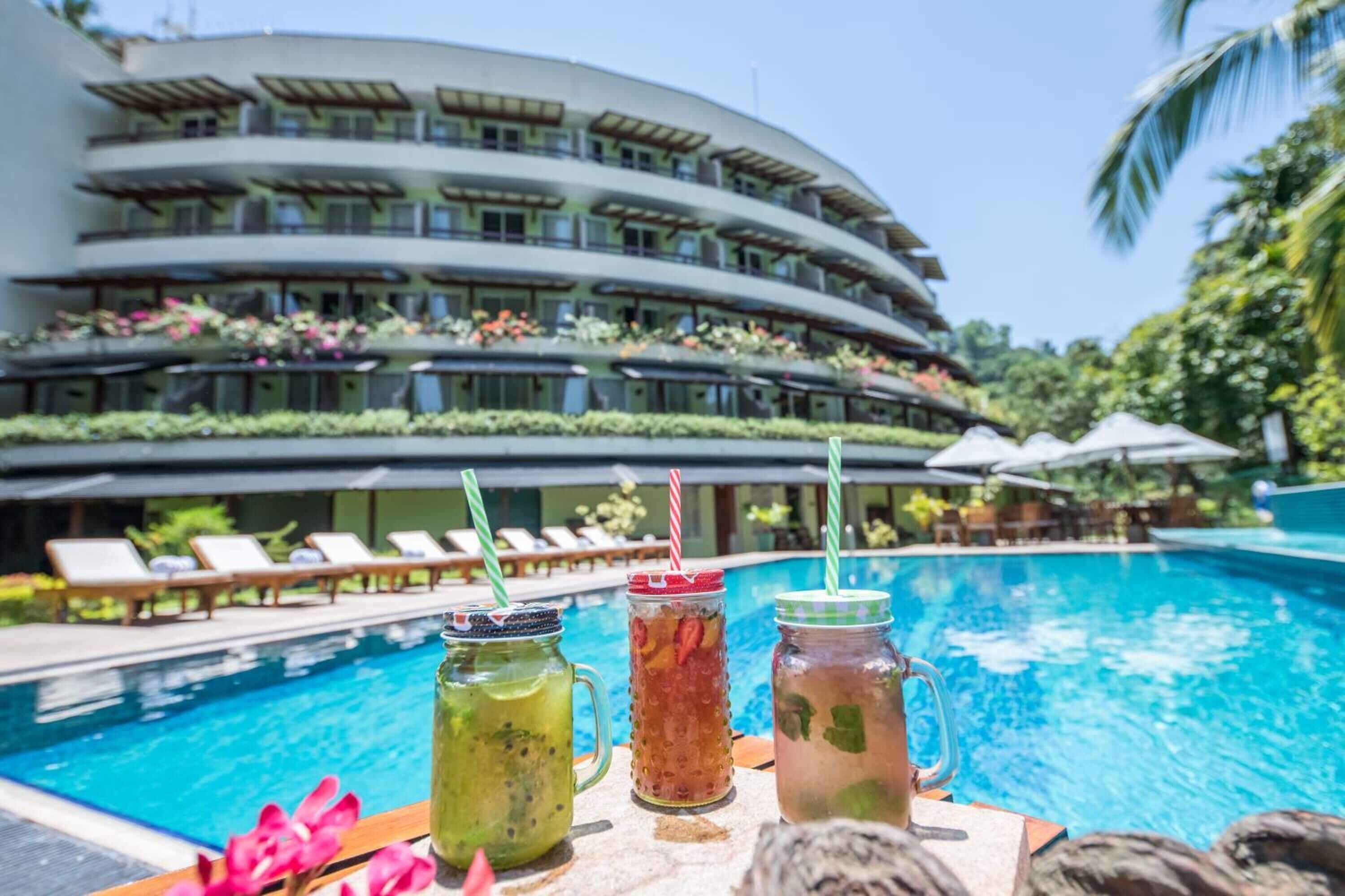 A poolside view with a modern hotel building, lounge chairs, and three drinks on a table.