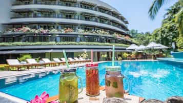 A poolside view with a modern hotel building, lounge chairs, and three drinks on a table.