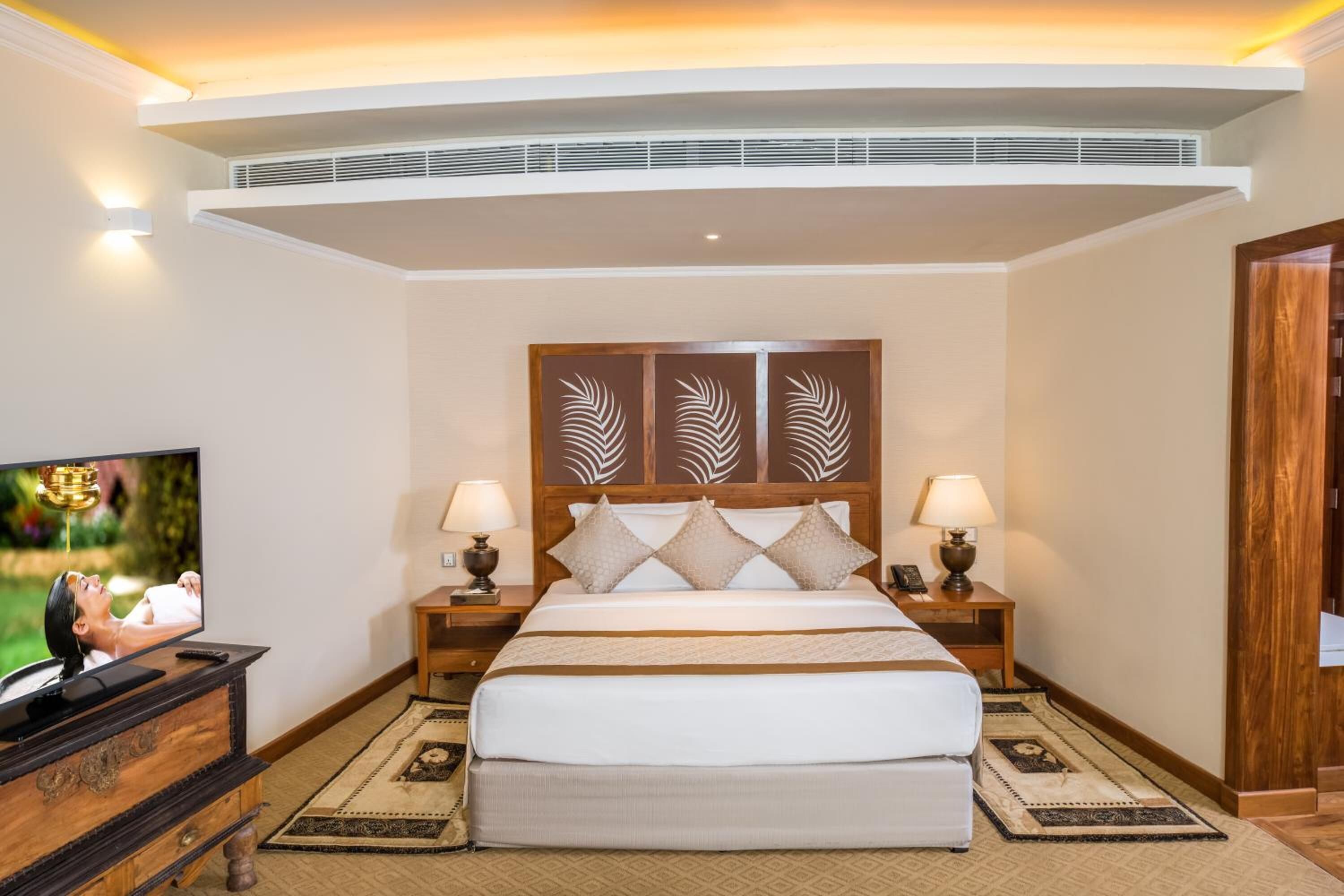 A hotel room with a large bed, two bedside tables with lamps, a flat-screen TV, a decorative headboard, and a ceiling vent.