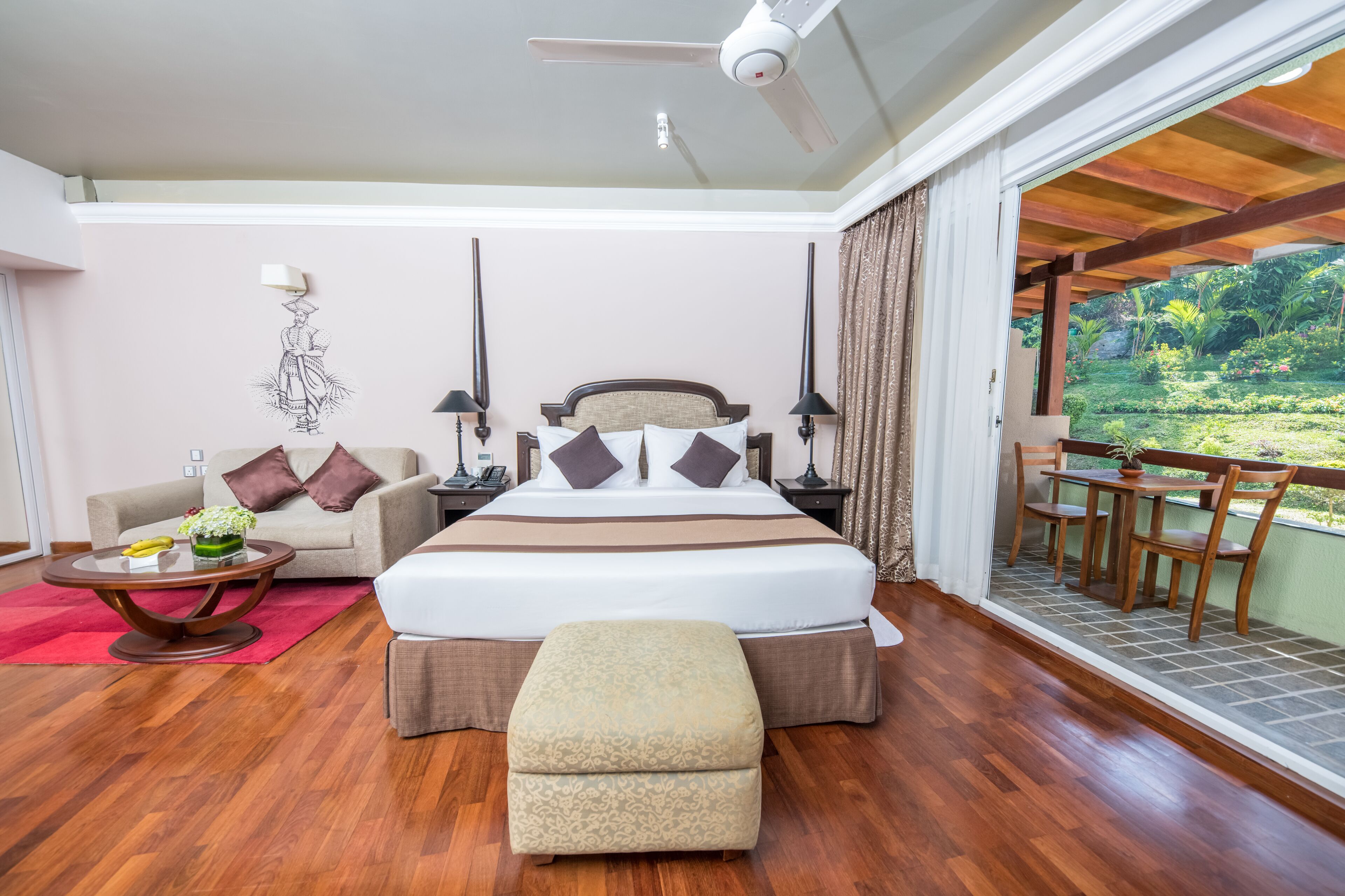 A spacious bedroom with a large bed, a sitting area with a sofa and a table, and a balcony with a view of greenery.