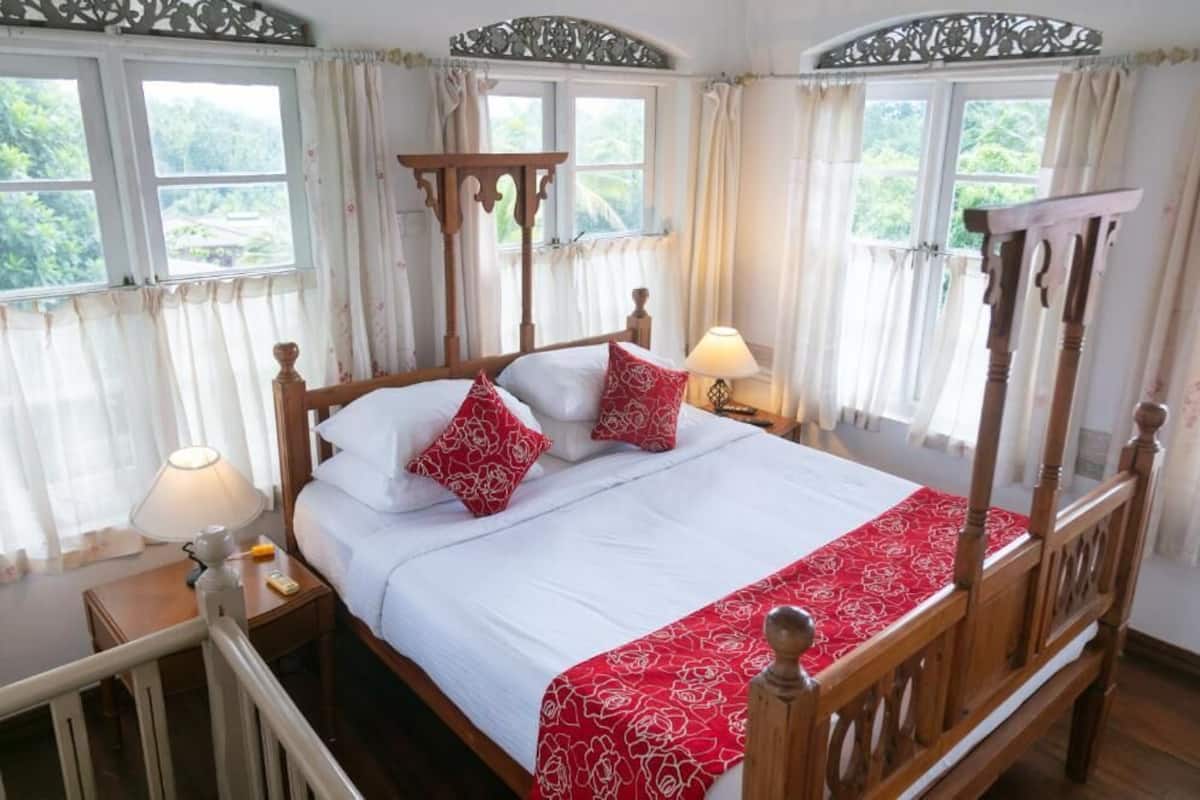 A four-poster bed with white bedding and red floral accents, two bedside tables with lamps, and a view of greenery through the windows.